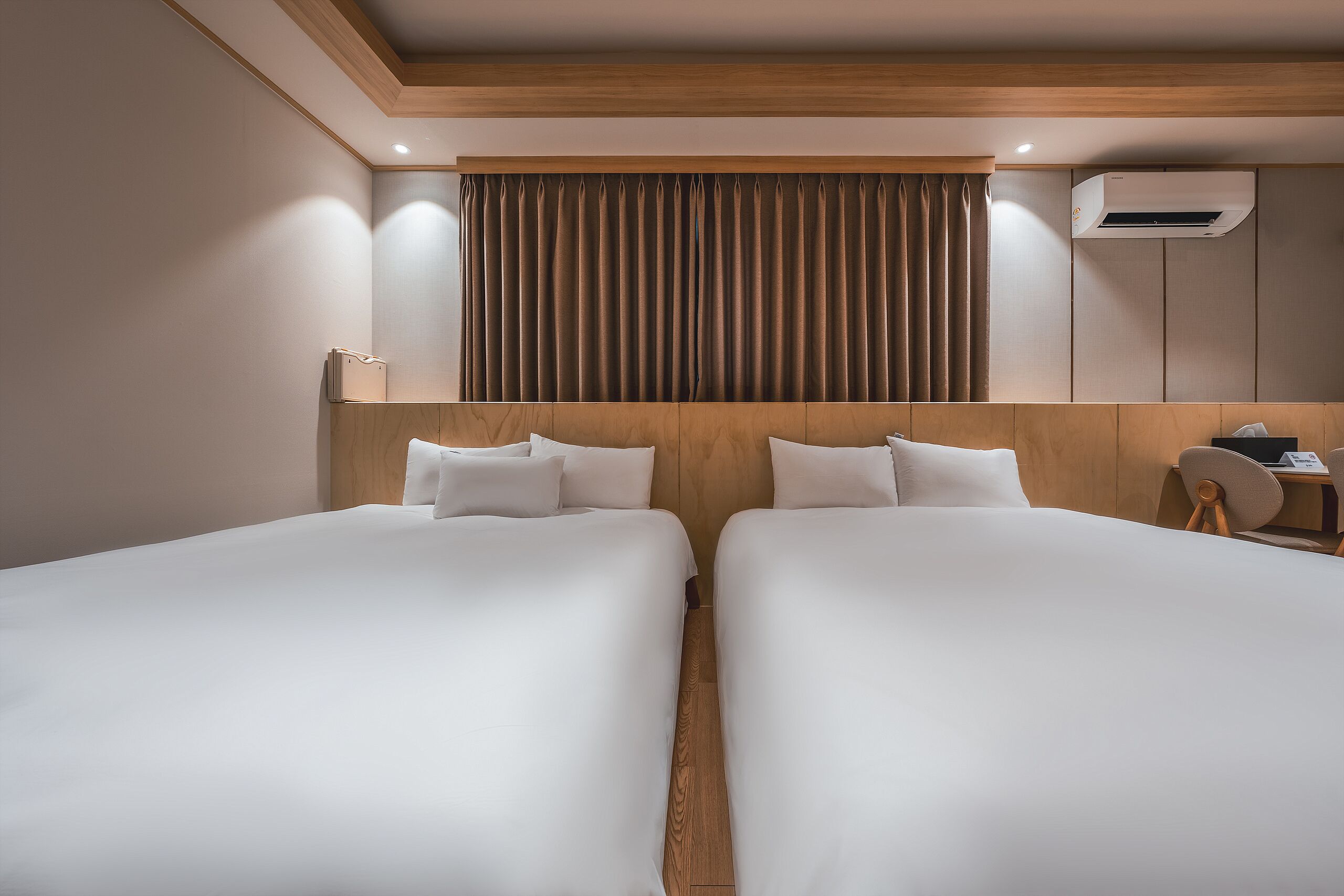 A hotel room with two beds, a wooden headboard, a window with curtains, and a wall-mounted air conditioner.