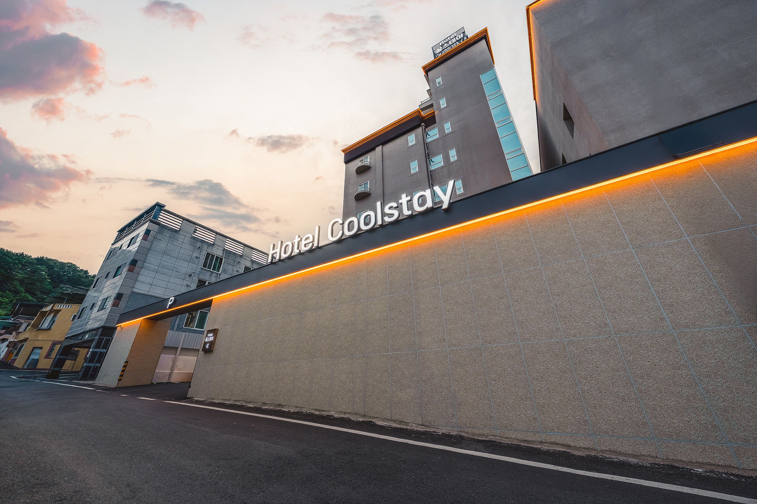 A hotel entrance with a modern design, featuring a prominent 'Hotel Coolstay' sign and a parking area.