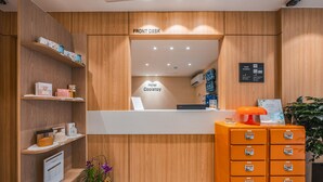 Área de recepção com acabamento em madeira, placa indicando 'Front Desk' e uma tela digital. Há prateleiras com diversos itens e uma planta.