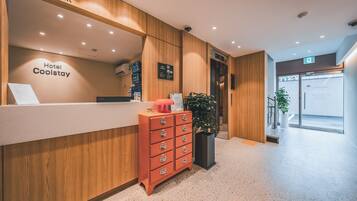 Un mostrador de recepción de hotel con detalles de madera, una barra para recepcionar a los huéspedes, un archivador rojo y una planta en maceta.