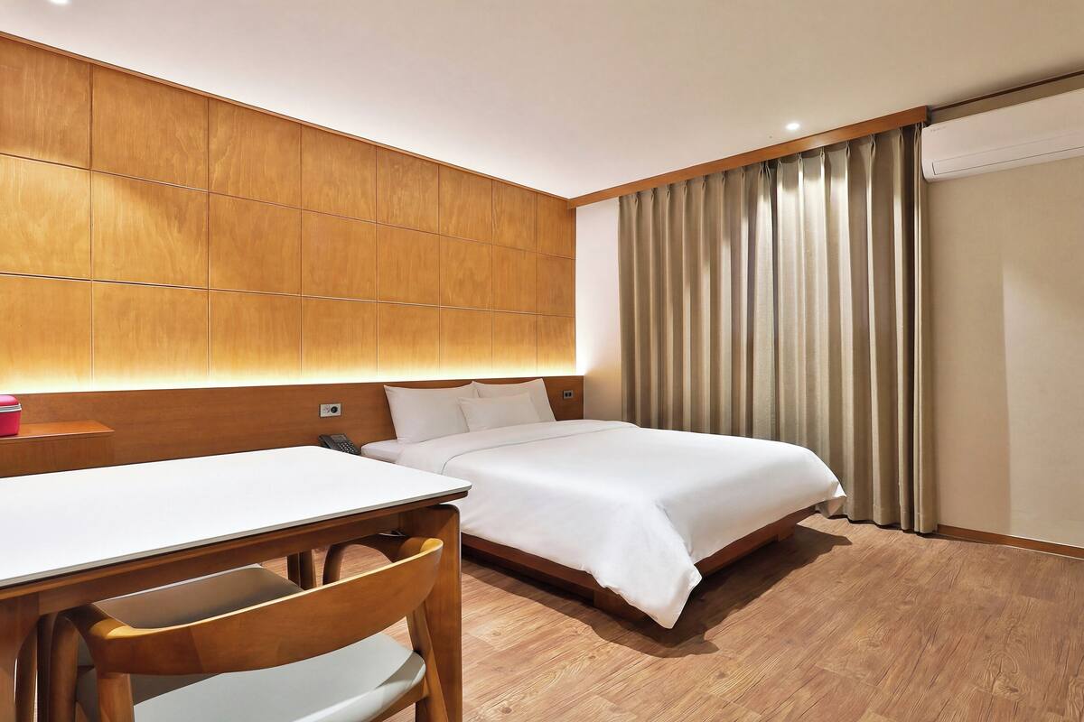 A hotel room with a wooden headboard, a bed with white bedding, a wooden desk with a chair, and a curtain on the window.