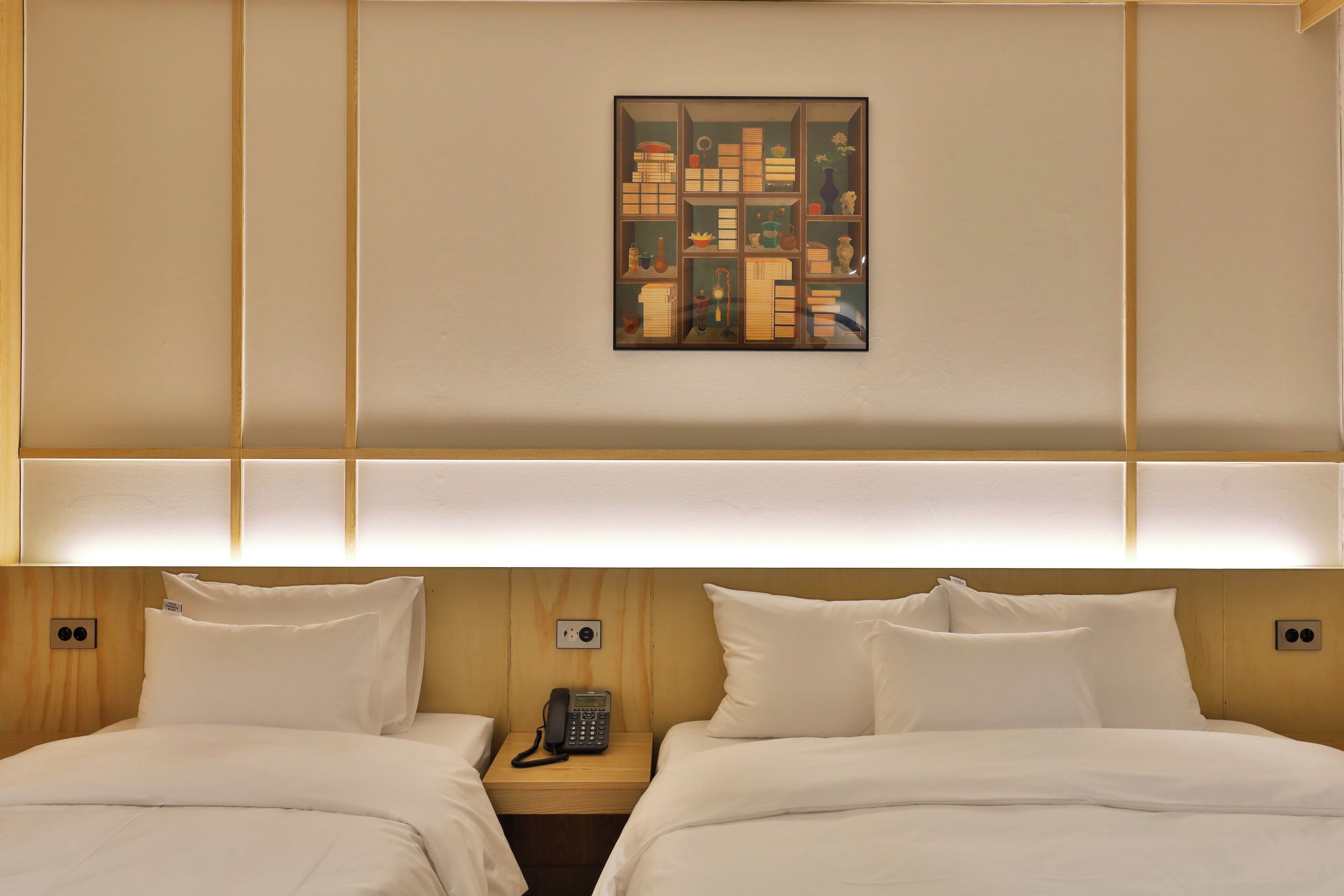 A hotel room with two beds, a bedside table with a phone, and a wall-mounted artwork.