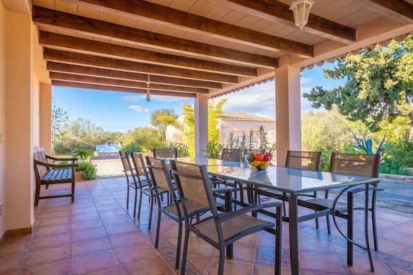 A covered patio with a glass table and chairs, surrounded by tiled flooring and wooden beams. - CAN SOMNI - Charming house 10 minutes from the beach with private pool and free WiFi (Ses Salines)