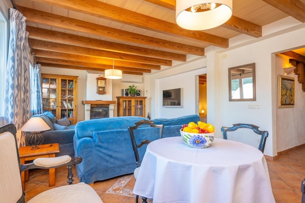 Ein gemütliches Wohnzimmer mit einem blauen Sofa, Holzbalken, einem Esstisch mit Obstschale und einem Kamin.