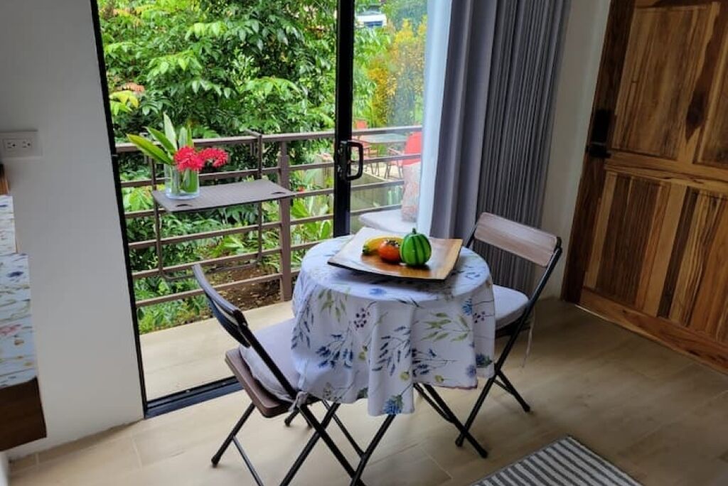 Un petit coin repas avec une table dressée pour deux personnes, une chaise et une vue sur un jardin luxuriant, visible à travers une porte vitrée.
