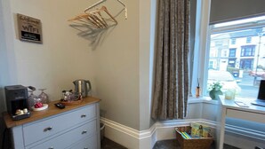 Habitación de hotel con escritorio, cafetera, hervidor, cesta con libros, ventana con cortinas y un tendedero de pared.