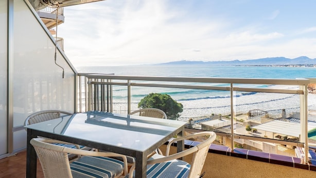 Ein Balkon mit Glastisch und Korbstühlen, der Blick auf einen Strand und den Ozean freigibt.