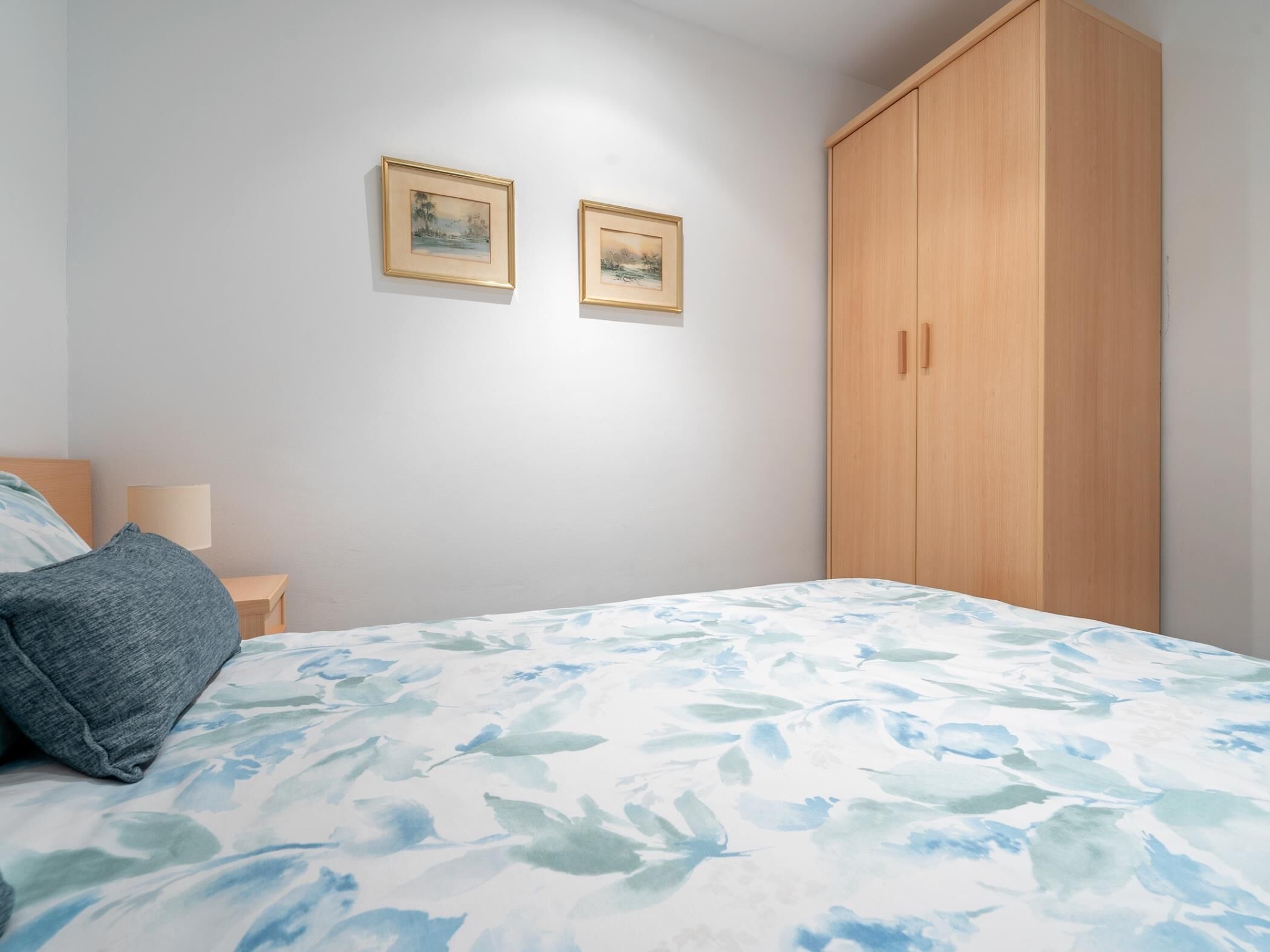 A bedroom with a bed, a nightstand, a wooden wardrobe, and two framed pictures on the wall.