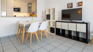 Ein modernes Wohnzimmer mit Flachbildfernseher, einem Essbereich mit weißem Tisch und Stühlen sowie einer Küche mit Mikrowelle und Schränken.