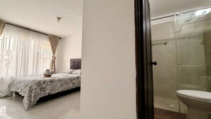 Un dormitorio con cama, una mesita de noche con toallas, una ventana con cortinas y una cabina de ducha de cristal.