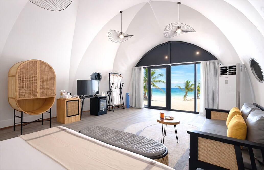 Ein modernes Hotelzimmer mit gewölbter Decke, einem großen Bett, einer Sitzecke mit Sofa und Sesseln, einem Fernseher, einem kleinen Tisch und Blick auf den Strand durch große Fenster.