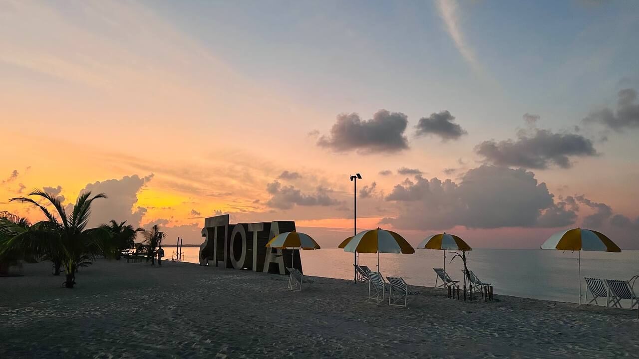 A beach with umbrellas and lounge chairs, a large sign with letters, and a sunset in the background.