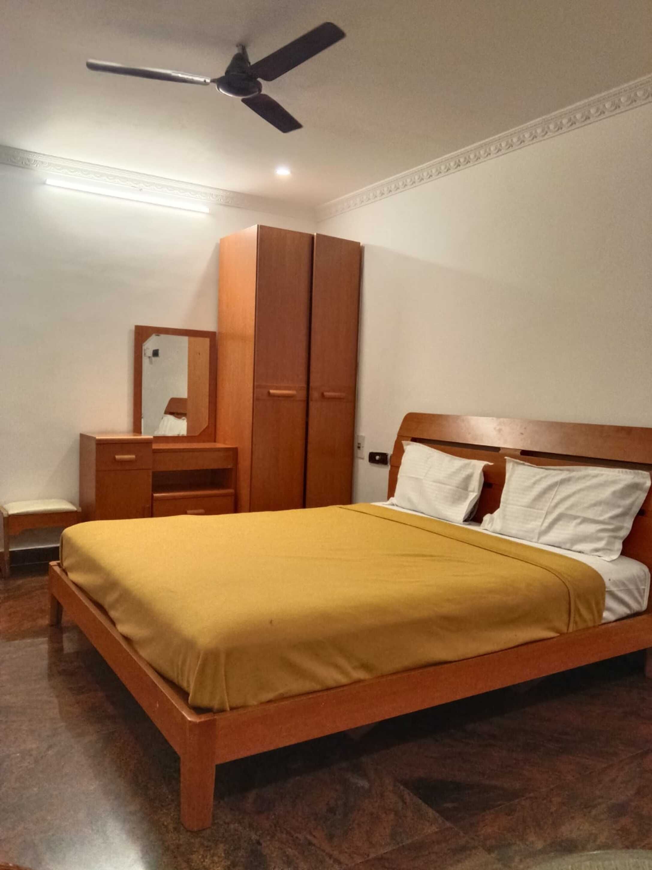 Ein Schlafzimmer mit einem Bett, einem Holzschrank, einem Schreibtisch mit Spiegel und einem Deckenventilator.
