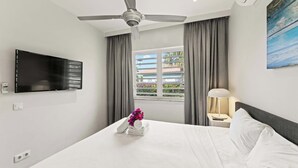 Habitación de hotel con cama, ventilador de techo, televisor montado en la pared, ventana con cortinas y un cuadro en la pared.