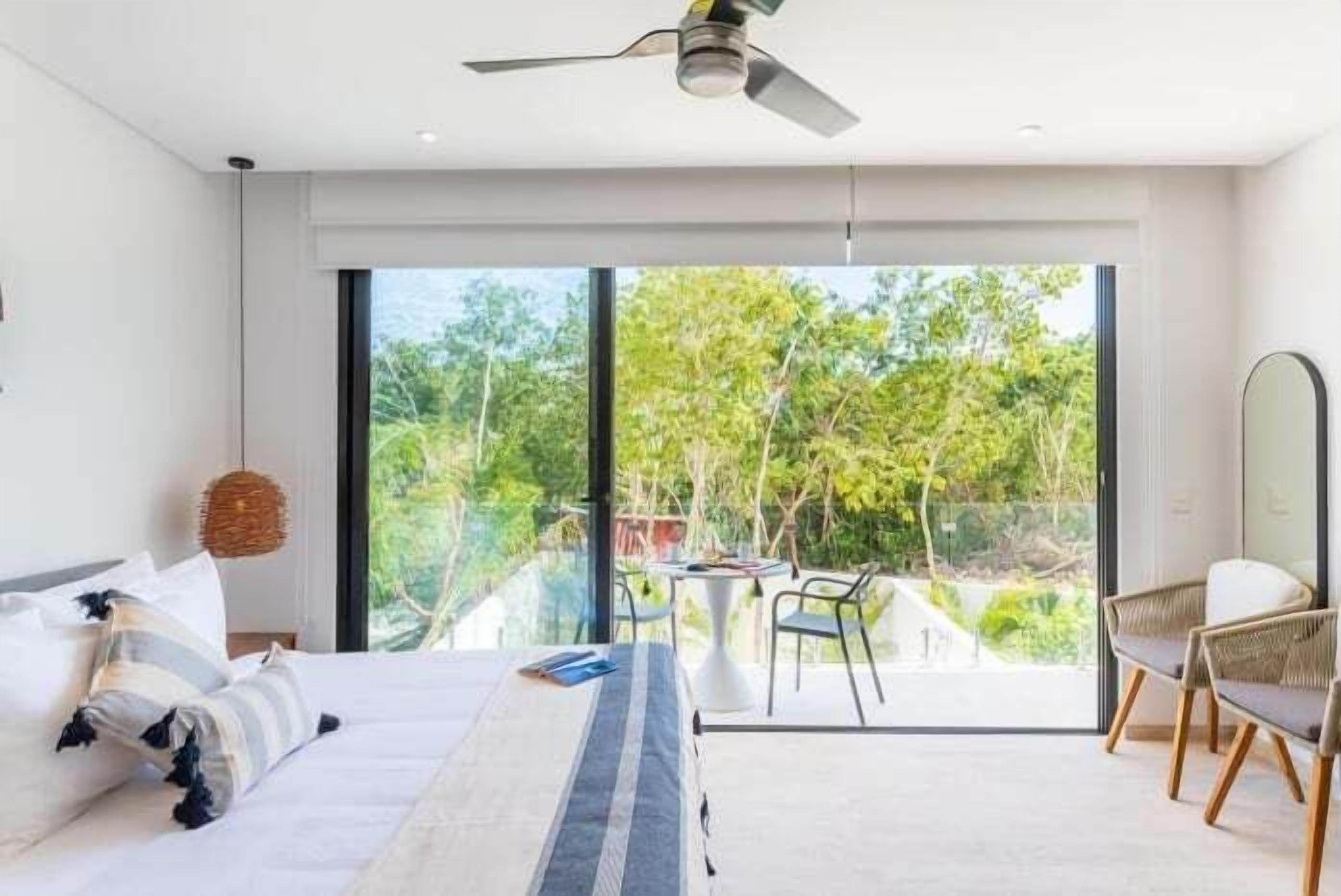 A modern bedroom with a large bed, a ceiling fan, and a view of greenery outside.