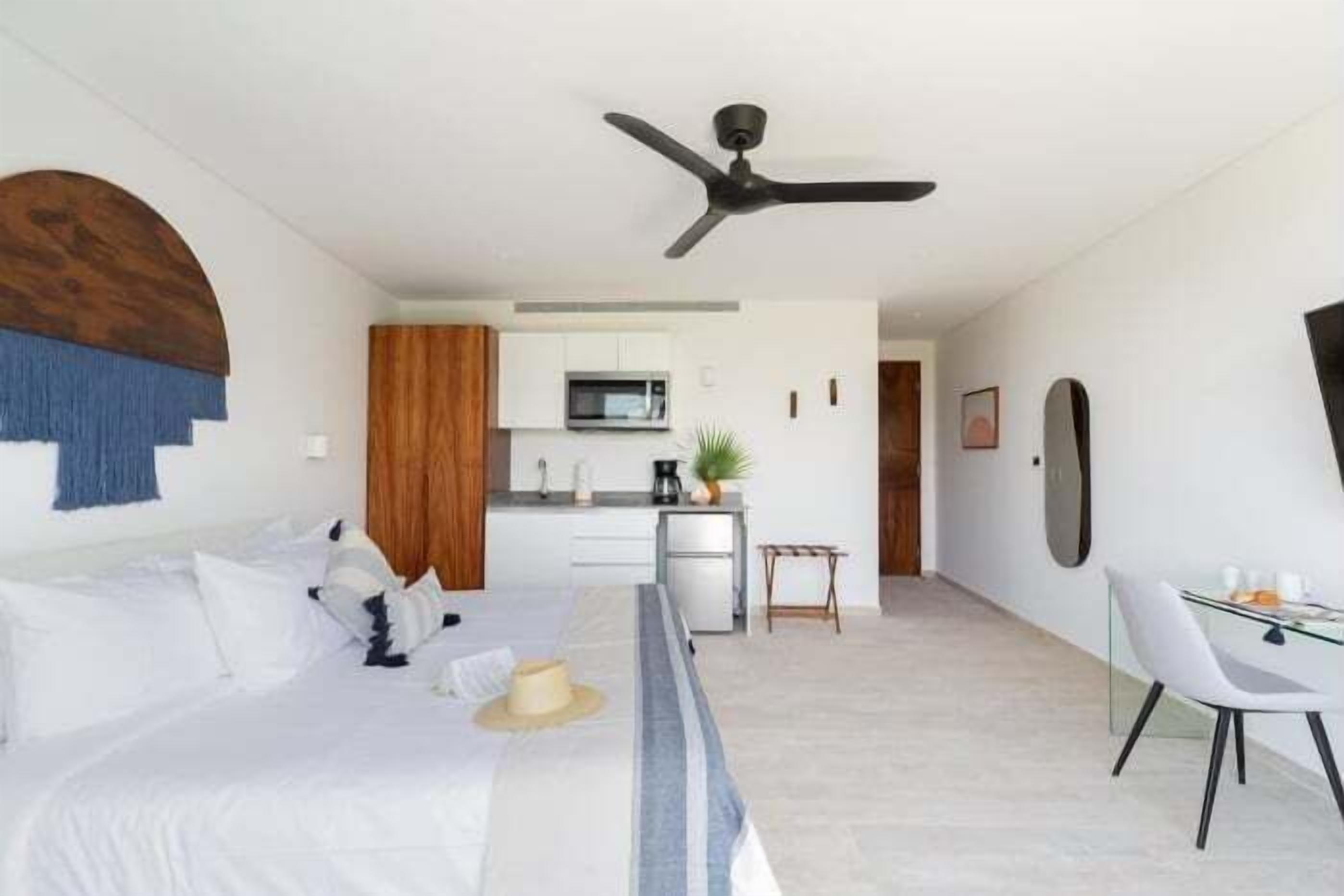 A modern hotel room with a bed, a desk, a chair, a ceiling fan, and a kitchenette.