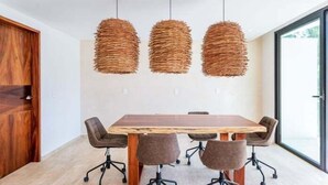 Uma sala de conferência com uma mesa de madeira e seis cadeiras, três luminárias pendentes de vime e uma grande janela.