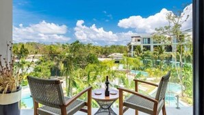 Un balcon agrémenté de fauteuils en osier et d’une table, d’où l’on peut admirer une piscine et une végétation luxuriante.