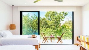 Un dormitorio moderno con una cama, una mesita pequeña y una silla, un ventilador de techo y un ventanal con vistas a zonas verdes.