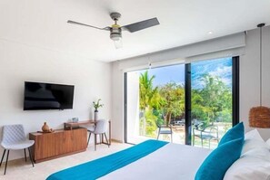 Ein modernes Schlafzimmer mit Bett, Fernseher, Schreibtisch und einem großen Fenster mit Blick auf Grünflächen.