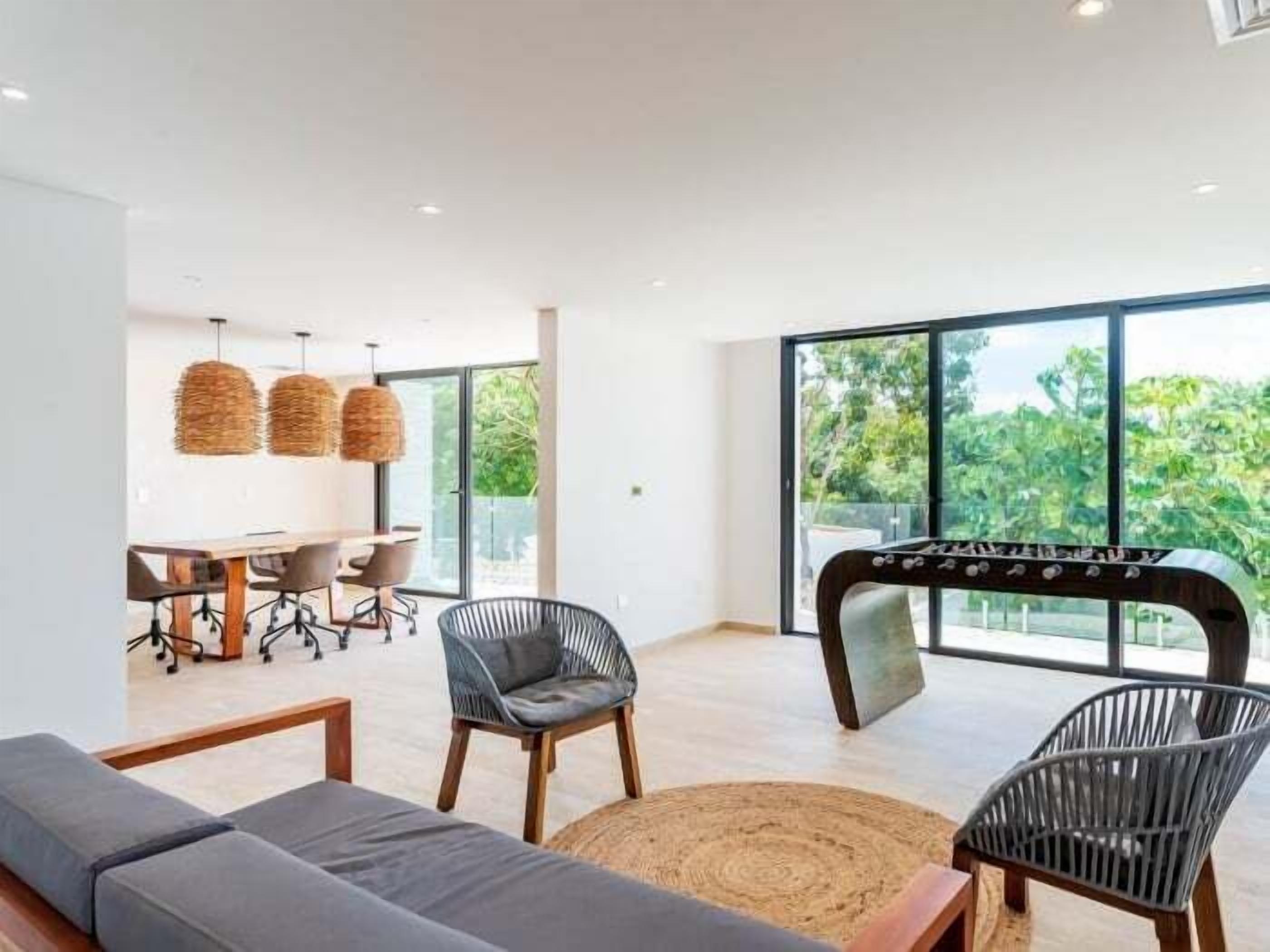 Un salon moderne avec un canapé, deux fauteuils, une table basse et un baby-foot. On aperçoit à l’arrière-plan un coin repas avec une table et des chaises.