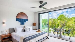 A bedroom with a large bed, a ceiling fan, a nightstand, a bedside table, and a view of greenery through a large window. - D303 CPL, Cálido estudio con aire acondicionado (Tulum)