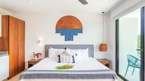 Una moderna camera d'albergo con un letto grande, una ventola a soffitto, un'opera d'arte appesa al muro, un comodino con un vaso di fiori e vista attraverso una porta vetrata.