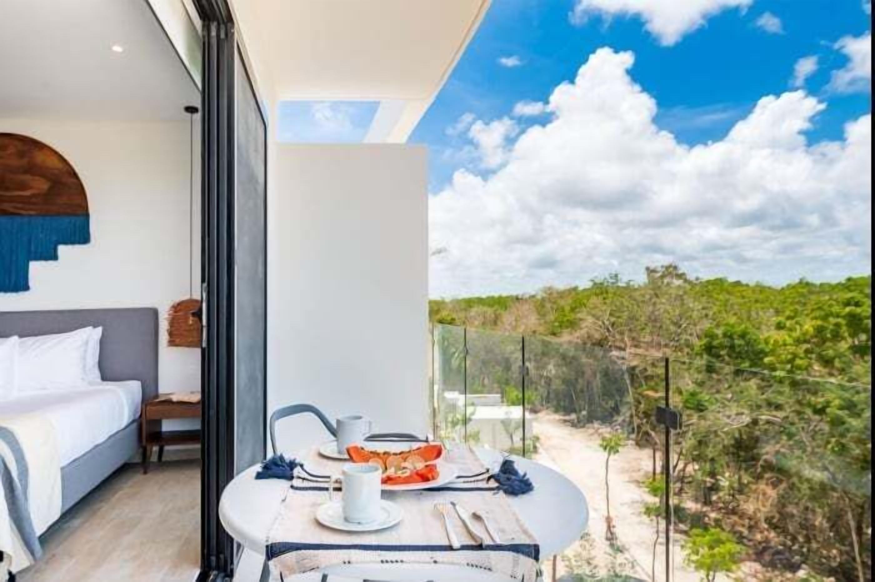 Ein modernes Zimmer mit Bett, Esstisch mit Frühstück und einer Glaswand mit Blick ins Freie.