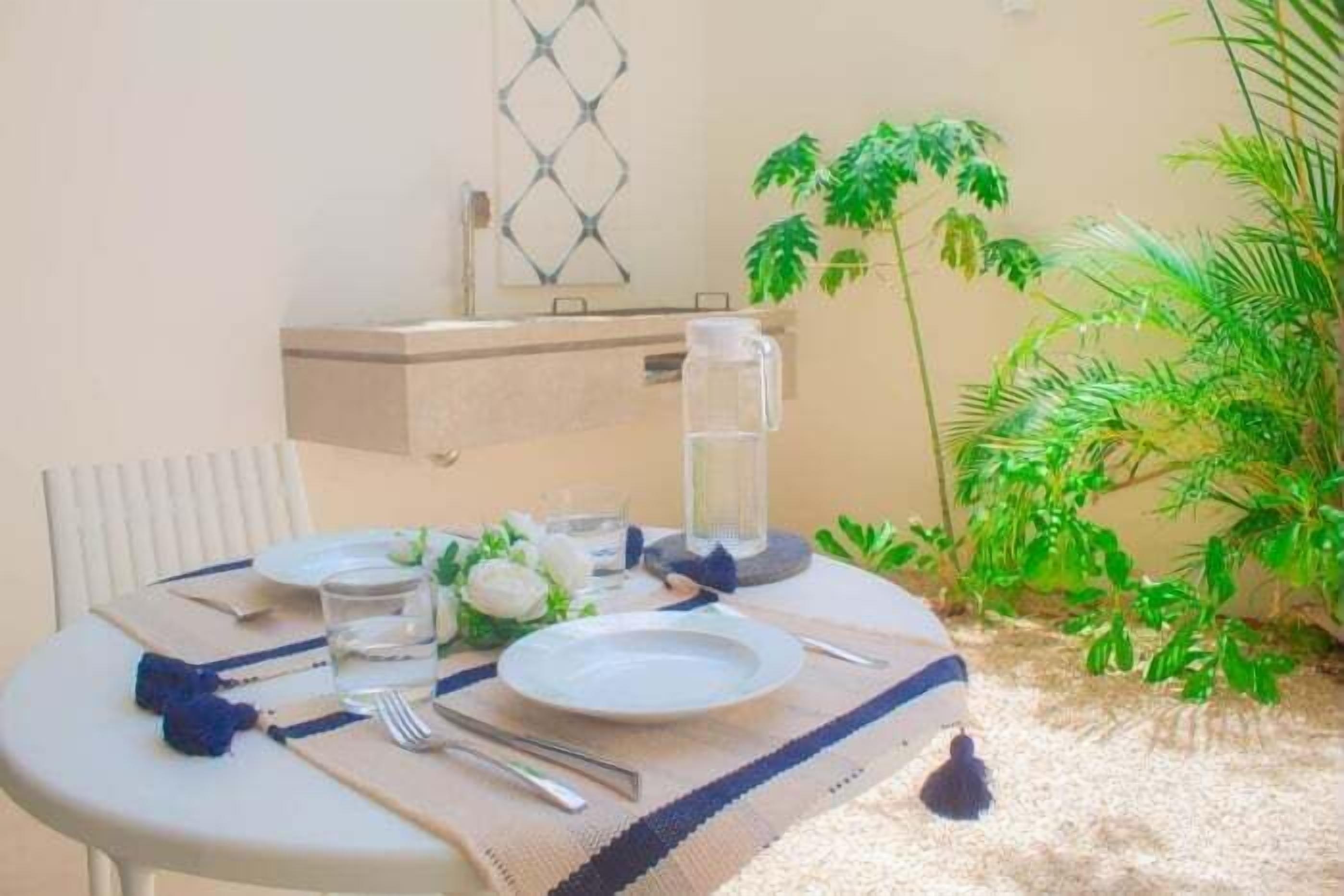 Mesa redonda para dos personas con mantel blanco, jarras de agua, platos, cubiertos y un arreglo floral decorativo.