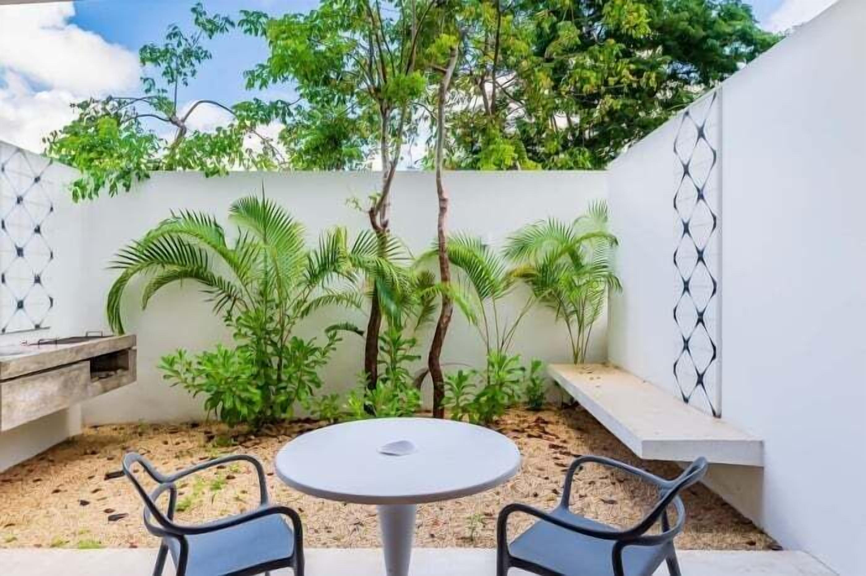 Eine Terrasse mit einem runden Tisch und zwei Stühlen, umgeben von üppiger Vegetation und einer weißen Wand mit geometrischem Muster.