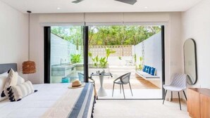 Une chambre moderne avec un lit, un ventilateur de plafond, une petite table avec des chaises et une vue sur un jardin, accessible par une porte coulissante.