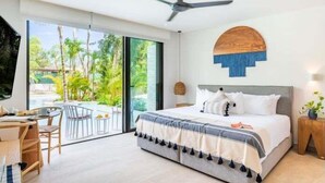 Camera da letto moderna con un letto, una ventola a soffitto, un'area pranzo con sedie e vista su una piscina e vegetazione.
