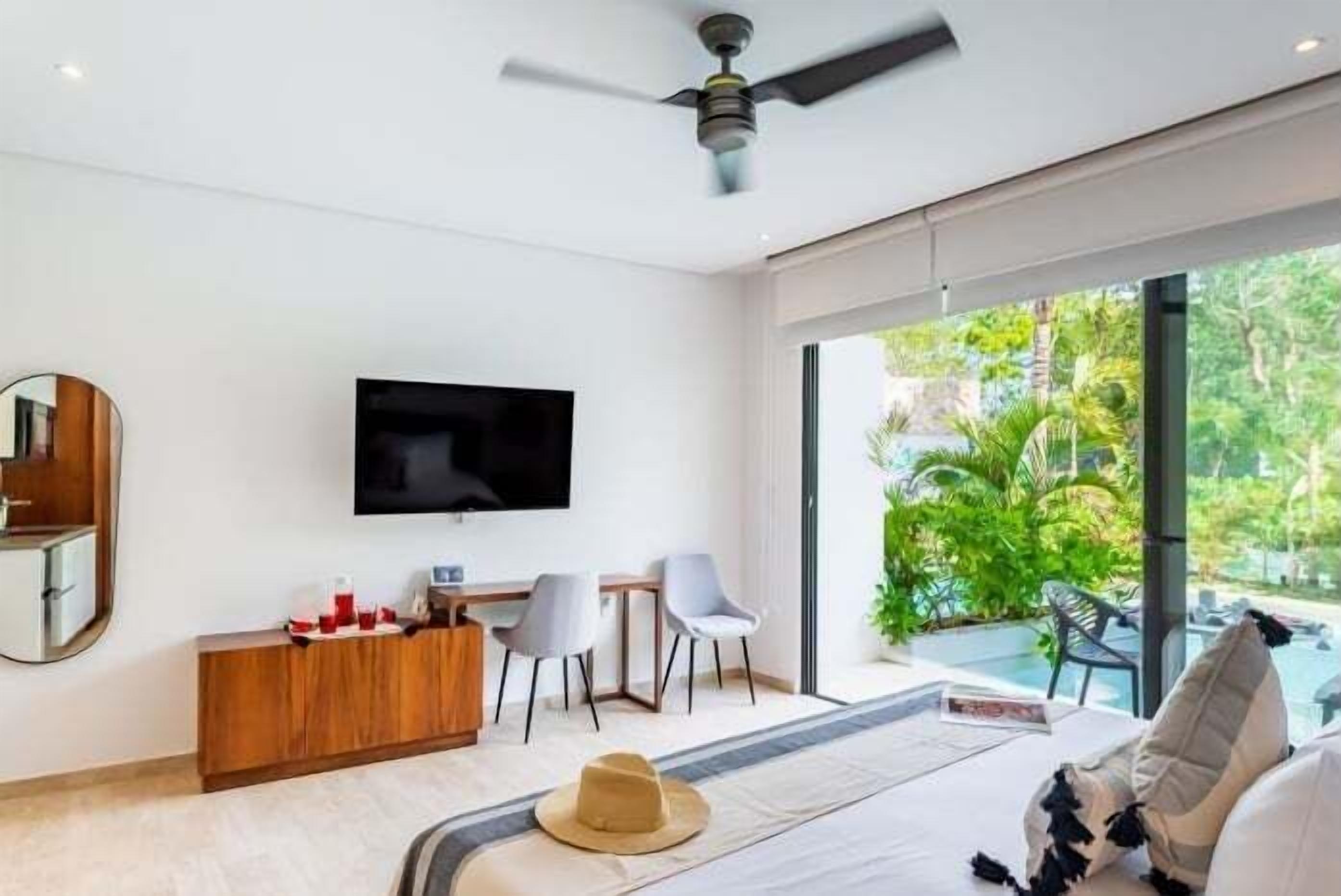A modern hotel room with a flat-screen TV, a wooden console, two chairs, a bed with a hat and pillows, and a large window with a view of greenery.