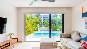 A modern living room with a TV, sofa, and a view of a swimming pool and greenery. - D104 CPL, Grandioso 2 bedrooms swim up (Tulum)