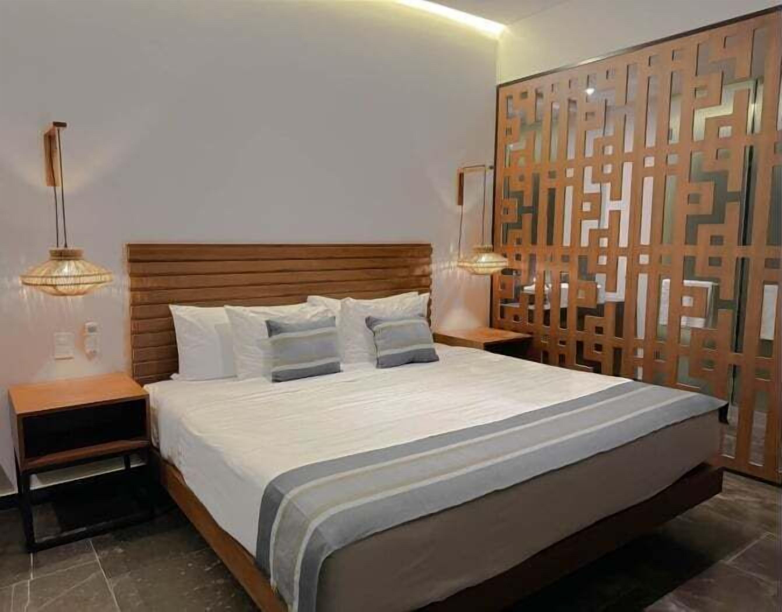 Quarto bem arrumado com cabeceira de madeira, cama grande com colcha listrada em cinza e branco, duas mesas de cabeceira com luminárias e um divisório decorativo de madeira.