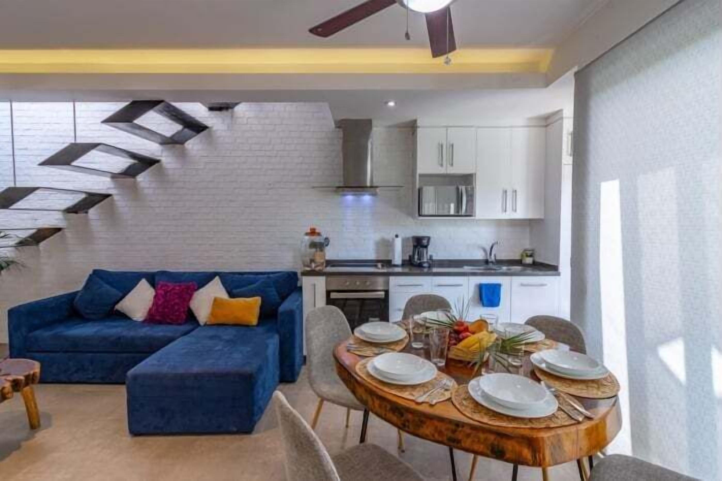 A modern living room with a blue sofa, a dining table set for four, and a kitchen in the background.