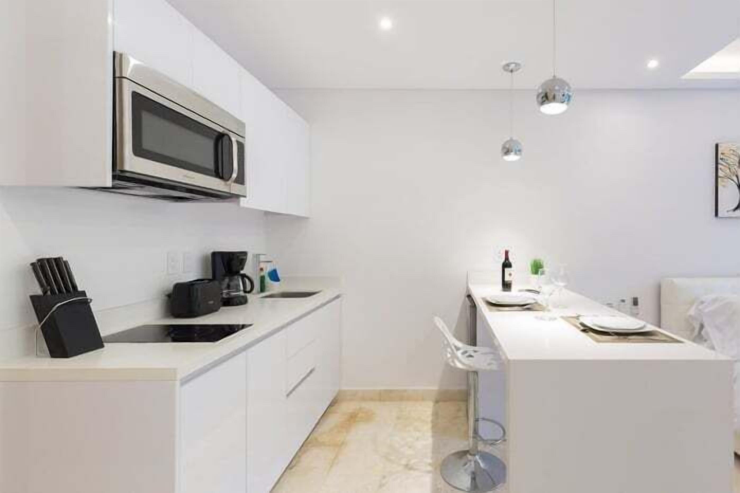 A modern kitchen with white cabinetry, a stainless steel microwave, a coffee machine, and a bar area with a wine bottle and glasses.