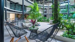 En atrium med flera våningsplan och modern arkitektur, med en central sittgrupp bestående av stolar och ett bord, omgiven av frodig grönska och växter.