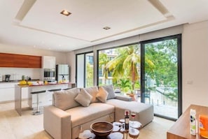 A modern living room with a sofa, a coffee table, and a view of a balcony with palm trees. - 205C PANOR, Large Apartment with 360° Pool (Tulum)