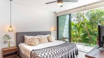 Une chambre à coucher comprenant un lit, des tables de chevet, un ventilateur de plafond et une grande fenêtre donnant sur un paysage verdoyant.
