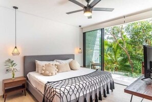 Ein Schlafzimmer mit Bett, Nachttischen, Deckenventilator und einem großen Fenster mit Blick ins Grüne.