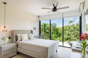 A modern bedroom with a bed, nightstands, a desk, and a large window offering a view of greenery. - PH8C PANOR, Luxury Penthouse Alberca Privada (Tulum)