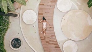 A person lying on a brick pathway surrounded by circular planters and large circular structures, with tropical foliage in the background. - PH8C PANOR, Luxury Penthouse Alberca Privada (Tulum)