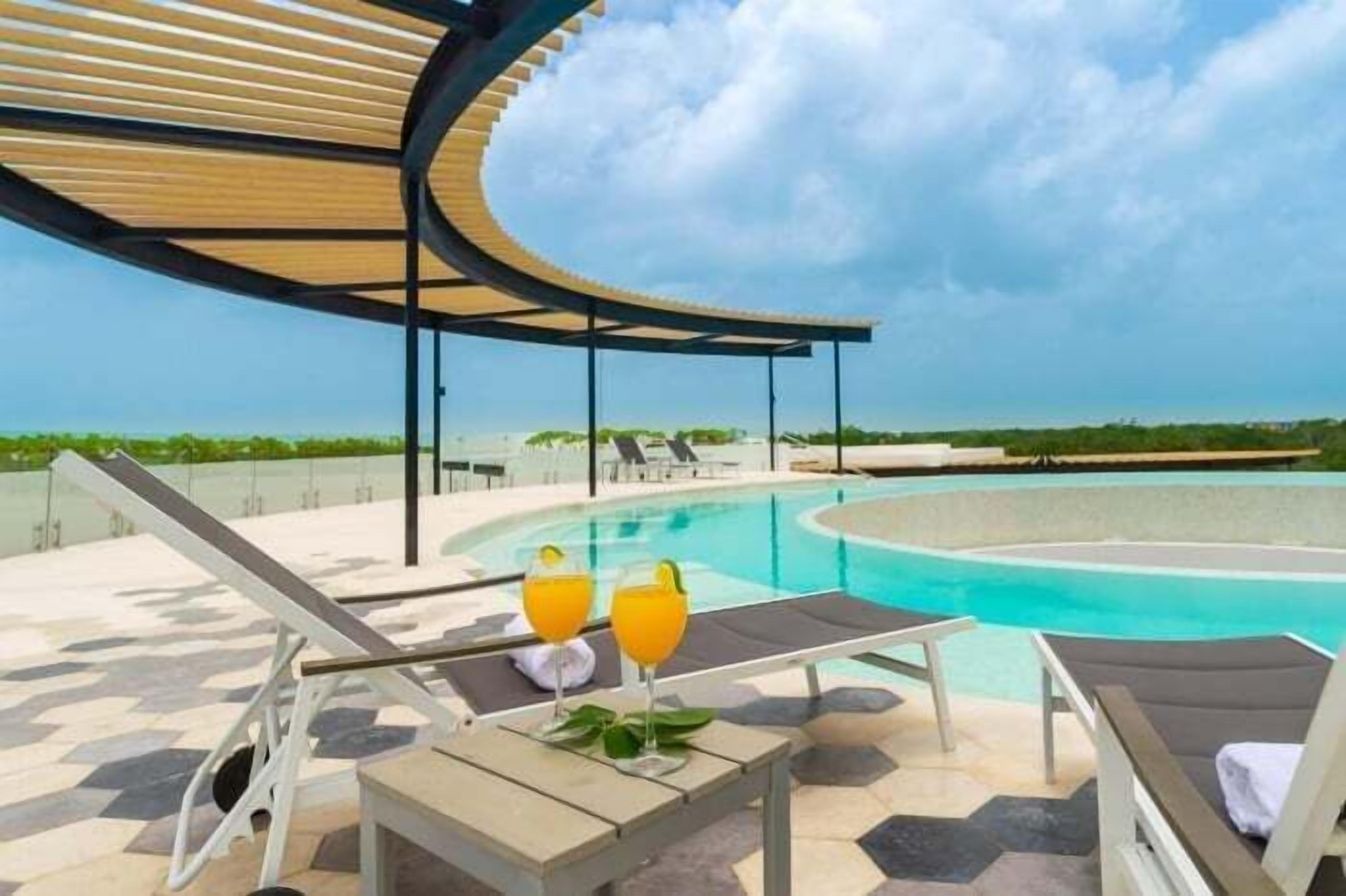Un espace piscine sur le toit, avec des chaises longues et une table, deux verres de jus d’orange et un auvent en bois incurvé.