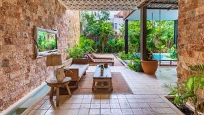 A covered patio area with stone walls, wooden furniture, and a view of a pool and greenery.