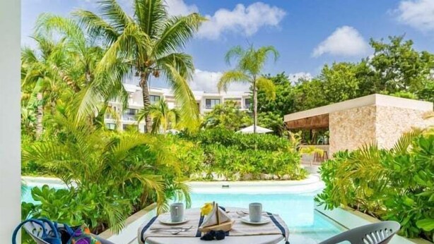 A poolside view with a table set for two, surrounded by lush greenery and palm trees, overlooking a building with multiple balconies. - A102 CPL, Warm Swim Up Studio-2 (Tulum)