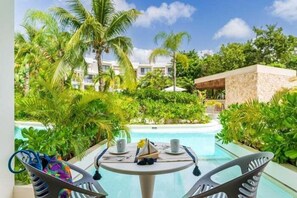 A poolside view with a table set for two, surrounded by lush greenery and palm trees, overlooking a building with multiple balconies. - A102 CPL, Warm Swim Up Studio-2 (Tulum)