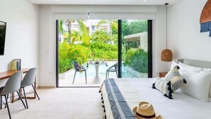 A modern bedroom with a bed, a desk, and a view of a lush garden through a sliding glass door. - A102 CPL, Warm Swim Up Studio-2 (Tulum)