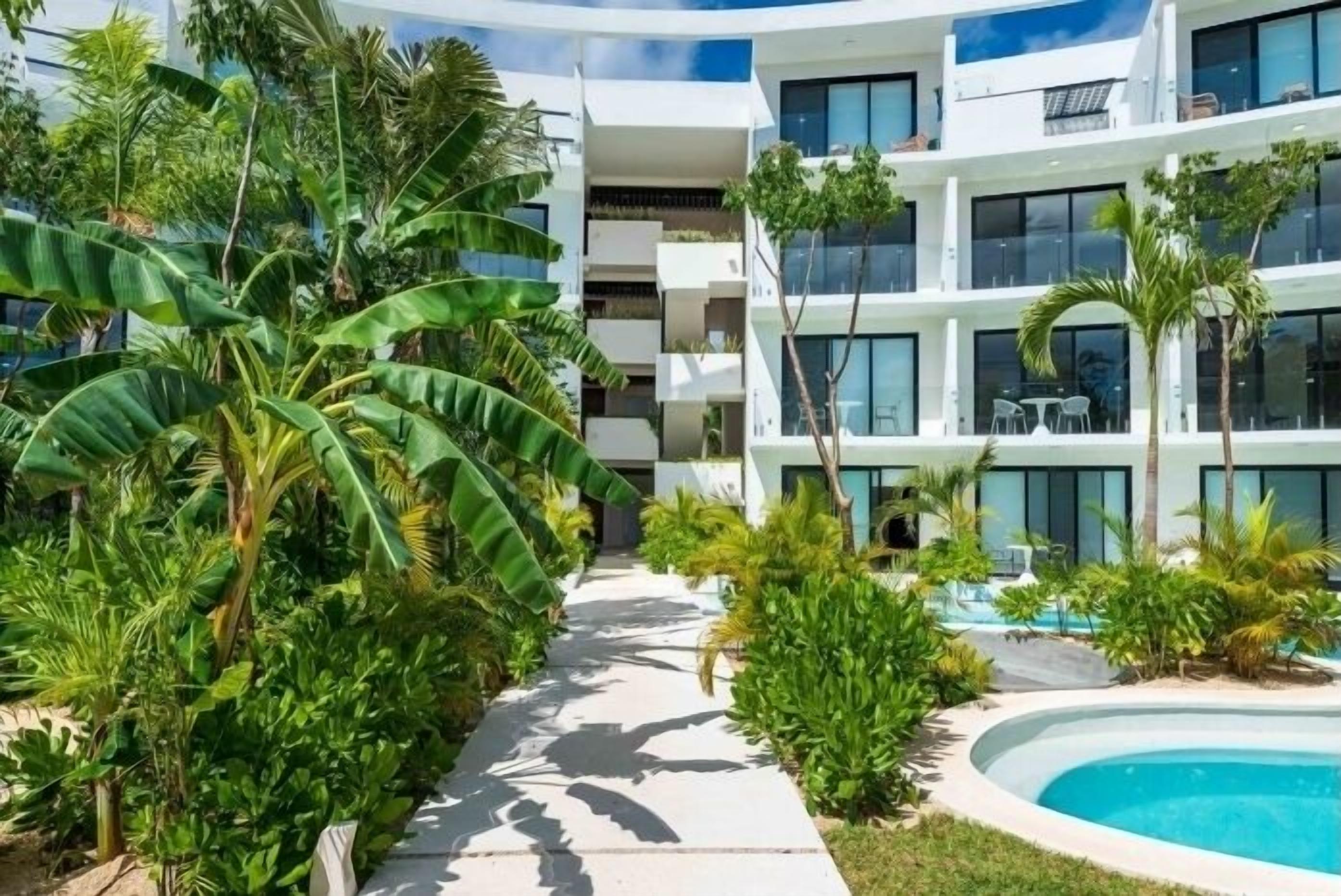 A modern hotel with a pool, surrounded by lush greenery and a clear sky.