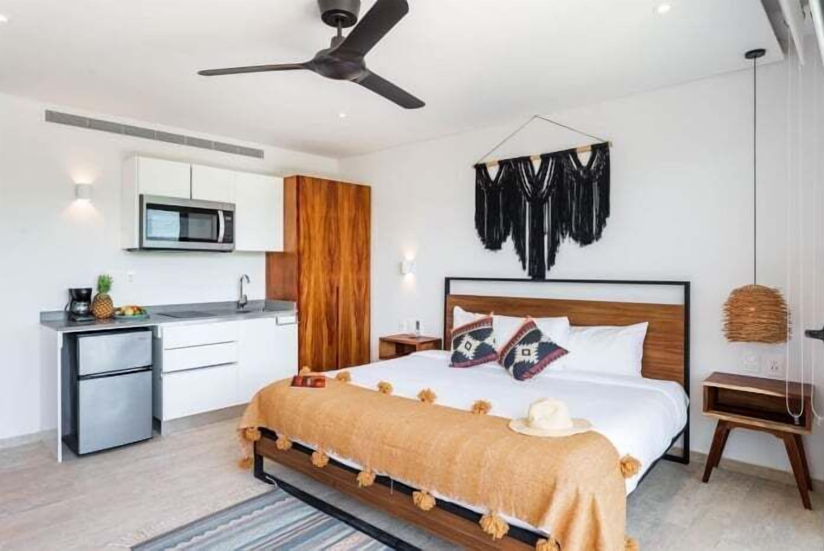 A modern hotel room with a bed, a small kitchenette, a ceiling fan, and a wall-mounted TV.